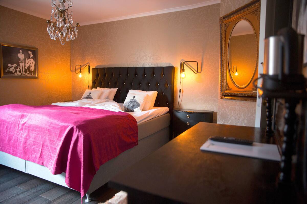 Ein ordentlich bezogenes Bett mit einer rosa Decke, ein Nachttisch mit einer Lampe und ein Spiegel an der Wand.