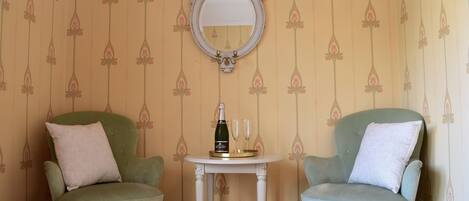 Two green armchairs with white cushions, a small white table with a bottle and glasses, and a mirror on the wall.