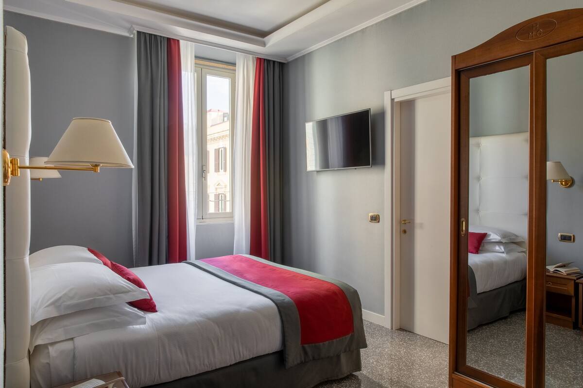 Una camera d'albergo con un letto, una televisione, uno specchio e una finestra con tende.