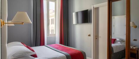 Habitación de hotel con cama, televisión, espejo y ventana con cortinas.