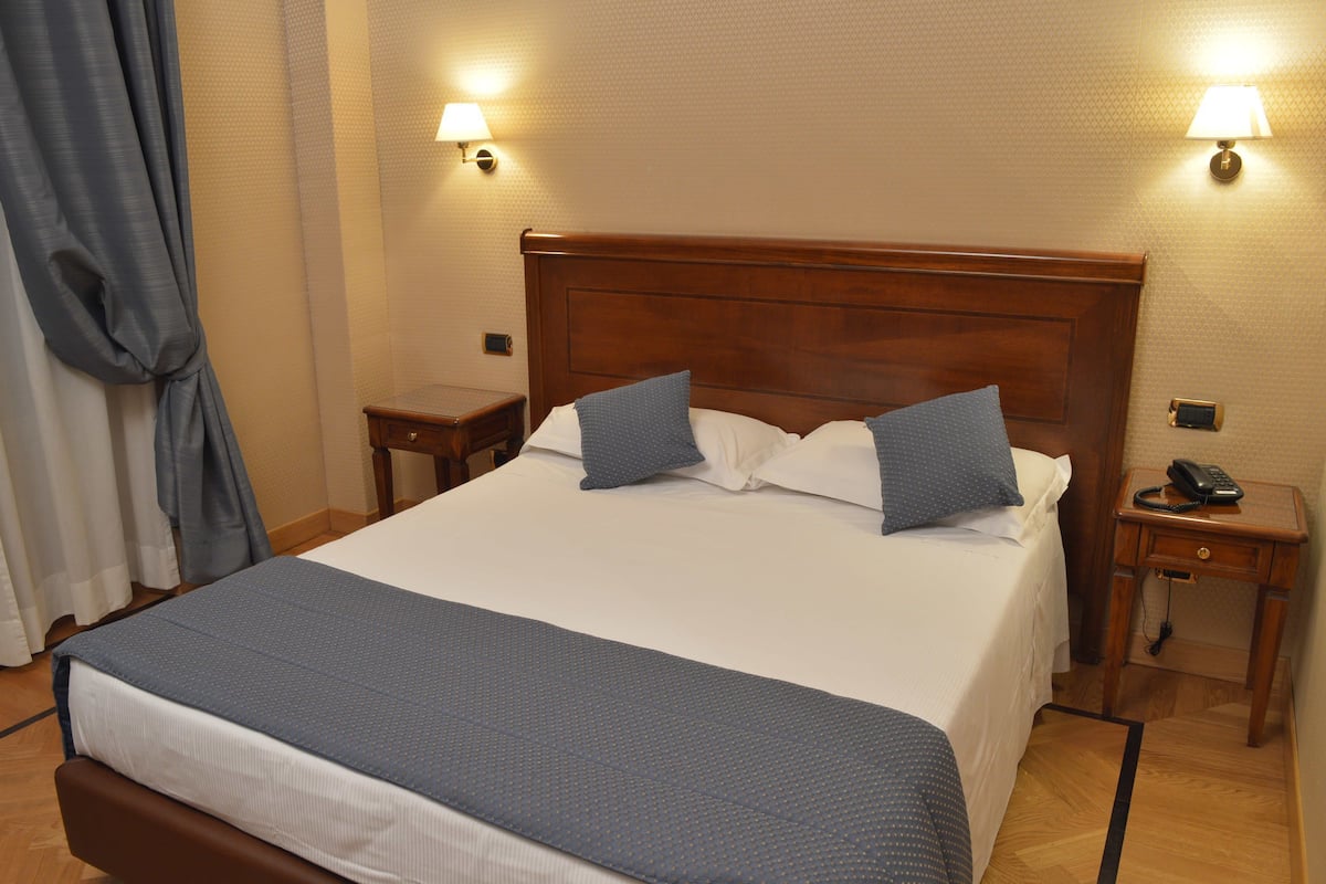 Un letto rifatto con cura, una testiera in legno, due comodini con lampade e una tenda blu parzialmente socchiusa.