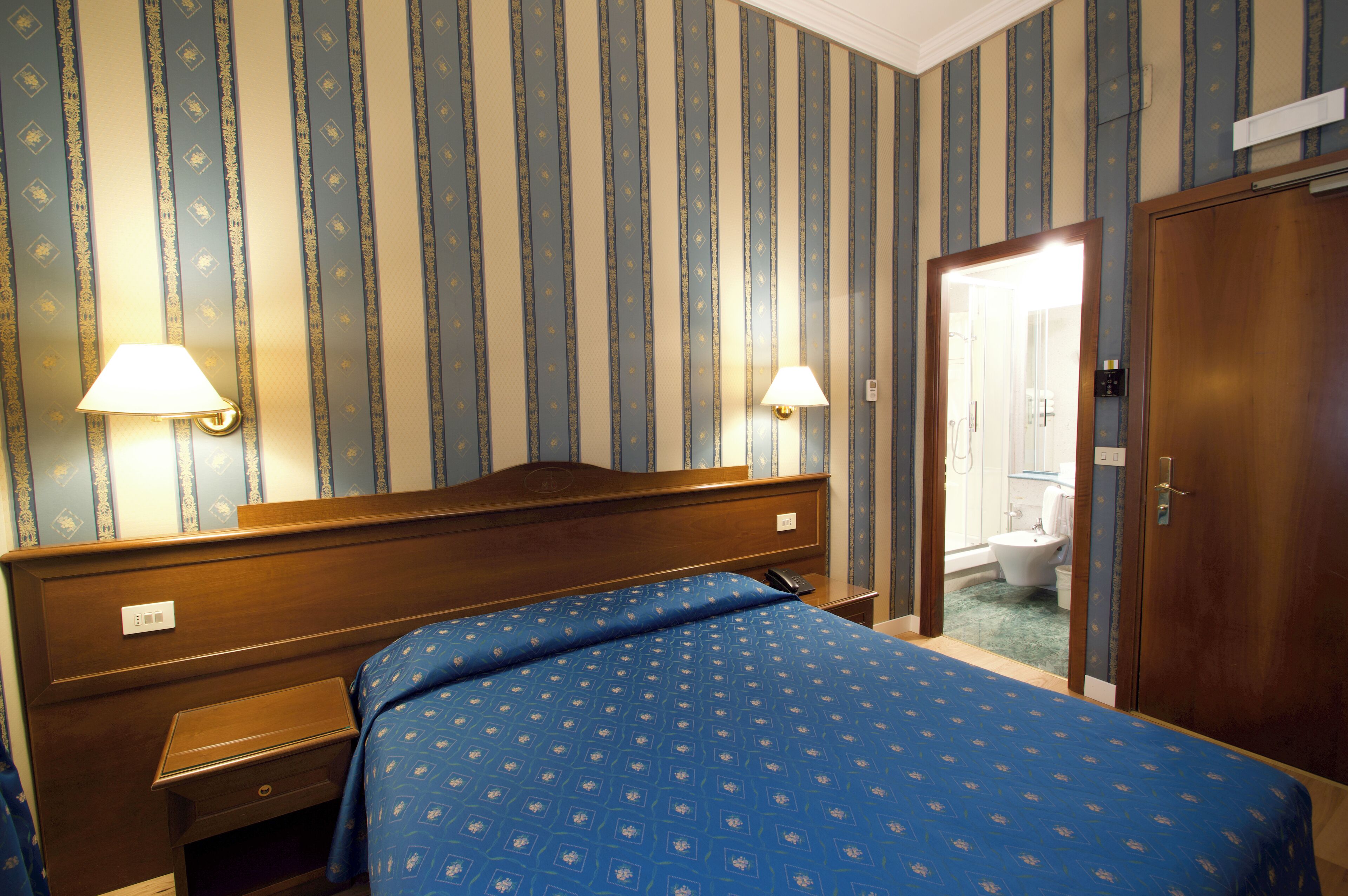 A hotel room with a wooden headboard, a blue patterned bedspread, two bedside lamps, and a door leading to a bathroom.