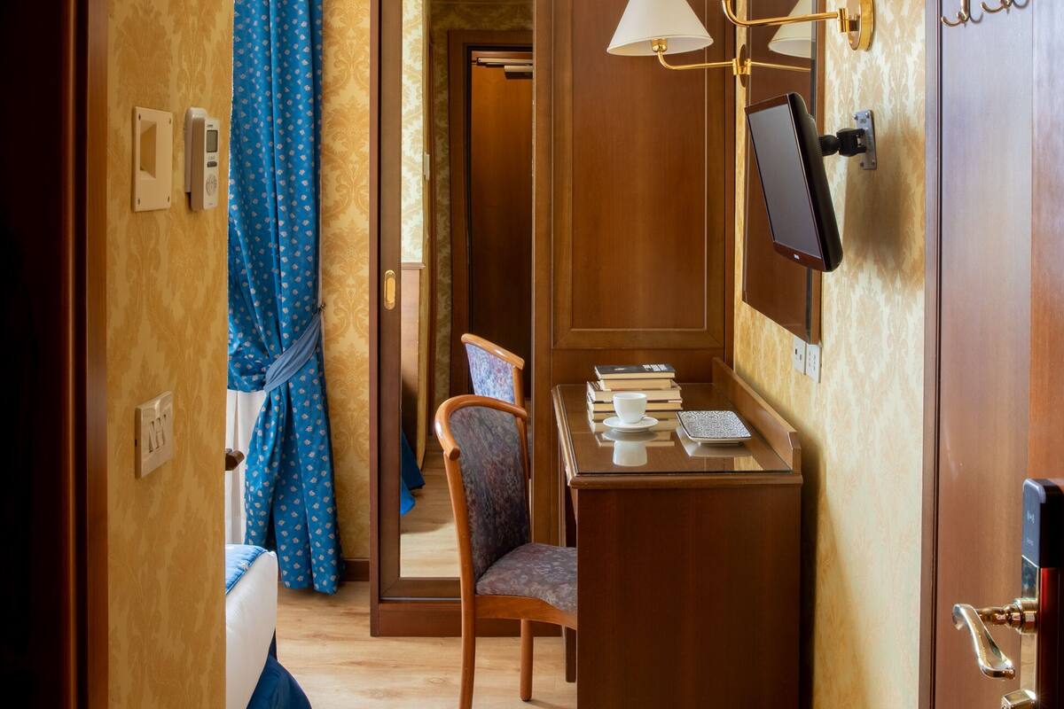 Una camera d'hotel con una scrivania in legno, una sedia, un letto con tende blu, una televisione a parete e una porta con uno specchio.