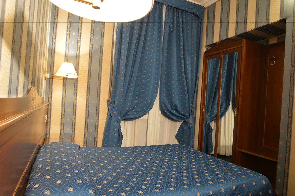 Una camera d'albergo con un letto, tende, un armadio in legno e una lampada.
