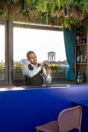 Un barman prépare un cocktail dans un bar offrant une vue sur un immeuble et un balcon.