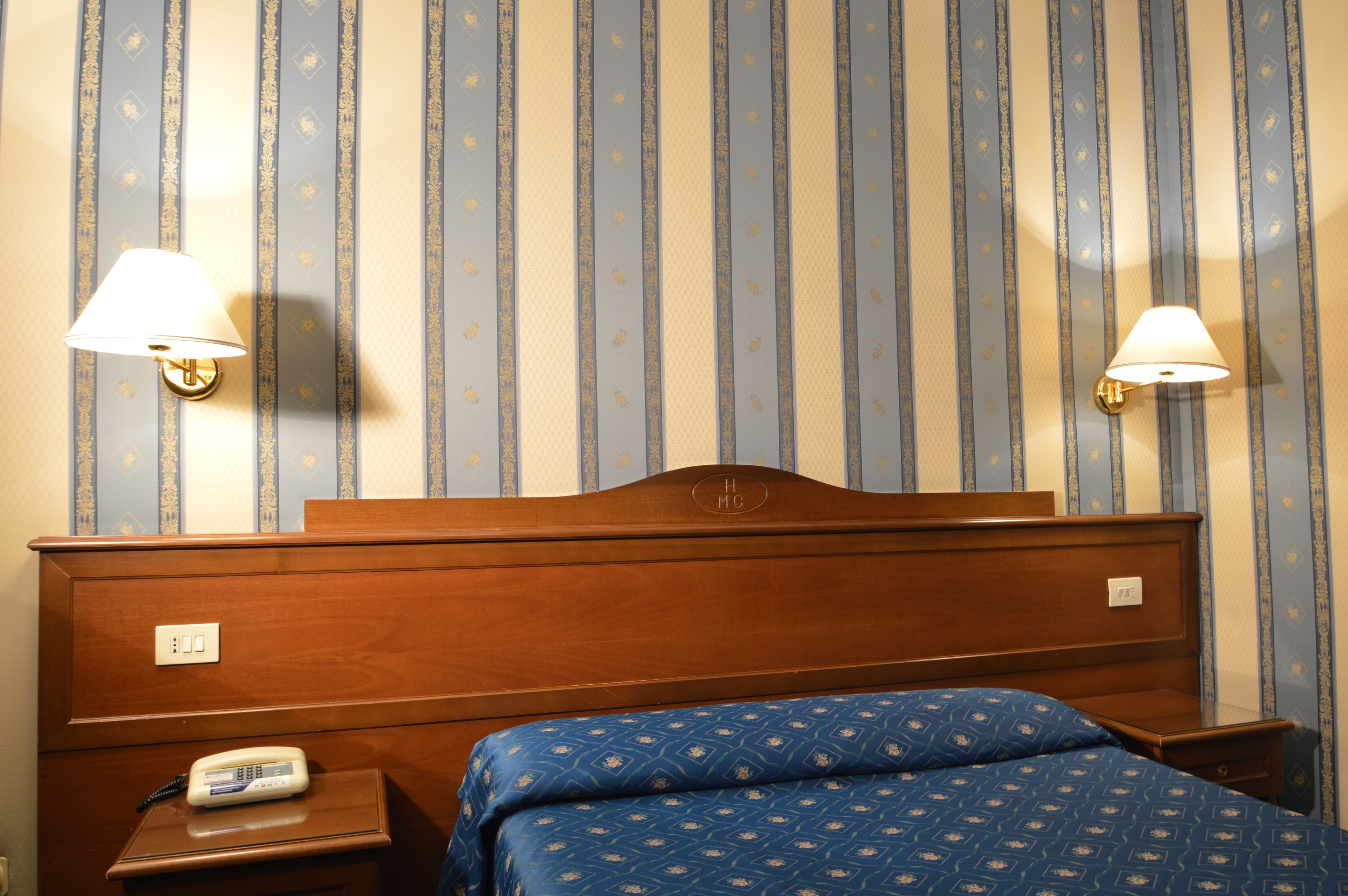 A hotel room with a wooden headboard, two bedside lamps, a bedside phone, and a bed with a patterned blue bedspread.