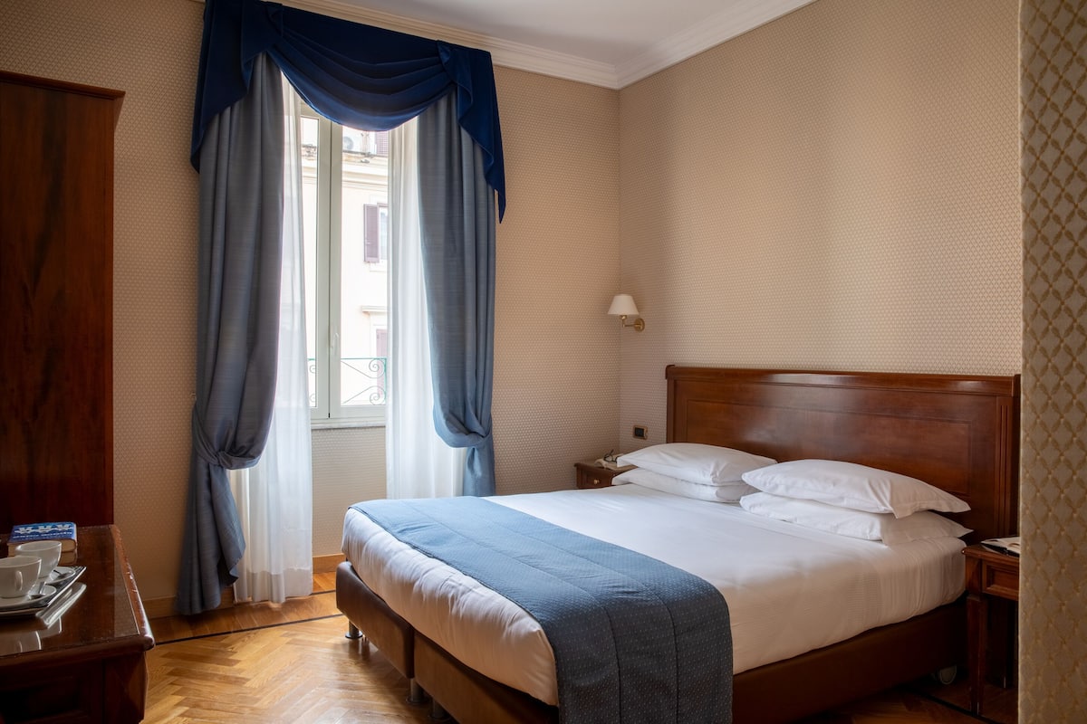 Un letto rifatto con lenzuola bianche, una testata in legno e una coperta blu. È presente un comodino in legno con una lampada e un telefono. La stanza ha un'ampia finestra con tende blu e vista su edifici esterni.
