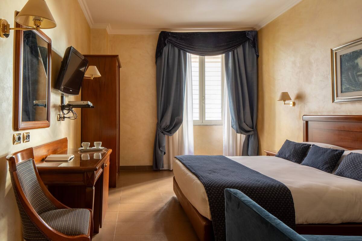 Una camera d'albergo con un letto, una scrivania con sedia, una televisione e una finestra con tende.