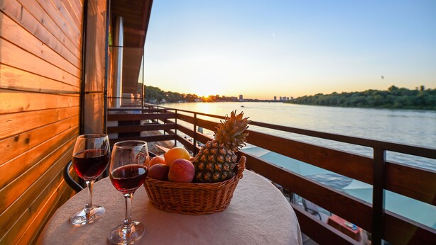 Une terrasse en bois avec une table sur laquelle se trouve un panier de fruits et des verres à vin, donnant sur une rivière avec un coucher de soleil en arrière-plan.