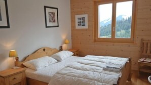 A wooden-paneled bedroom with a large bed, two bedside lamps, and a window with a mountain view. - Appartement Esserts A33 (Val d'Illiez)