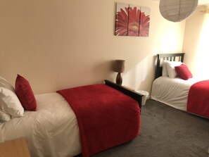 A hotel room with two beds, each with white bedding and red throw blankets. There is a wall-mounted artwork of a red flower, a bedside lamp, and a ceiling fan. - Three Bedroom House in Basildon (Basildon)