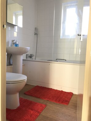 A bathroom with a toilet, sink, and shower. There are two red rugs on the floor. - Three Bedroom House in Basildon (Basildon)