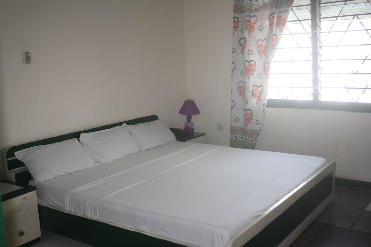 A single-bed room with a green headboard, white bedding, a nightstand with a lamp, and a window with patterned curtains.