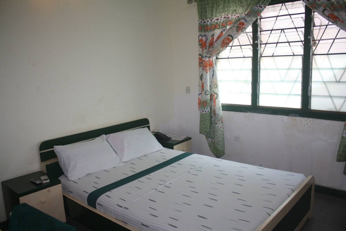 A single bed with a green headboard and white bedding, a nightstand with a phone, and a window with a patterned curtain.