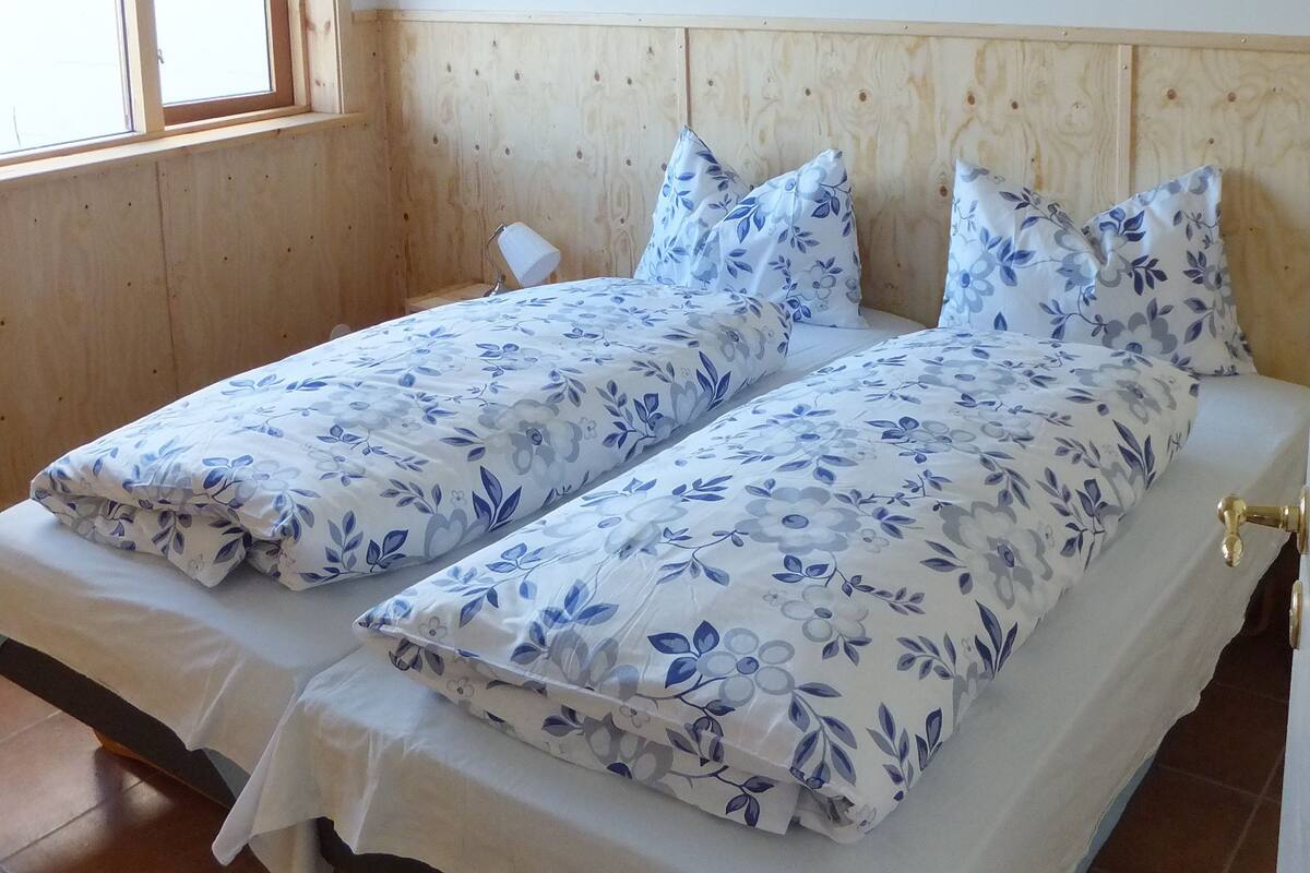 Huoneessa on kaksi erillistä sänkyä, puinen pääty, valkoiset seinät ja ikkuna, jossa on sälekaihtimet.