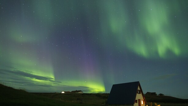 A house with a dark roof and a lit window, surrounded by a grassy landscape under a night sky with visible stars and the Northern Lights. - Brekkulækur Guesthouse (Hvammstangi)