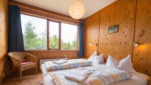 A wooden-paneled room with two beds, a wicker chair, and a large window with curtains. - Brekkulækur Guesthouse (Hvammstangi)