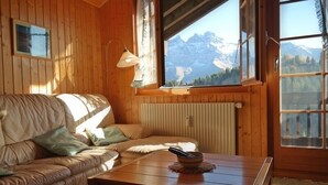 A cozy room with a leather sofa, a wooden table with a bowl, and a view of mountains through large windows. - Appartement Myrtilles 25 (Val d'Illiez)