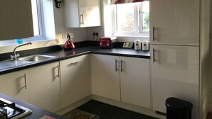 A compact kitchen with white cabinets, a black countertop, a red kettle, and a window with floral curtains. - Two Bedroom Apartment in Basildon (Basildon)