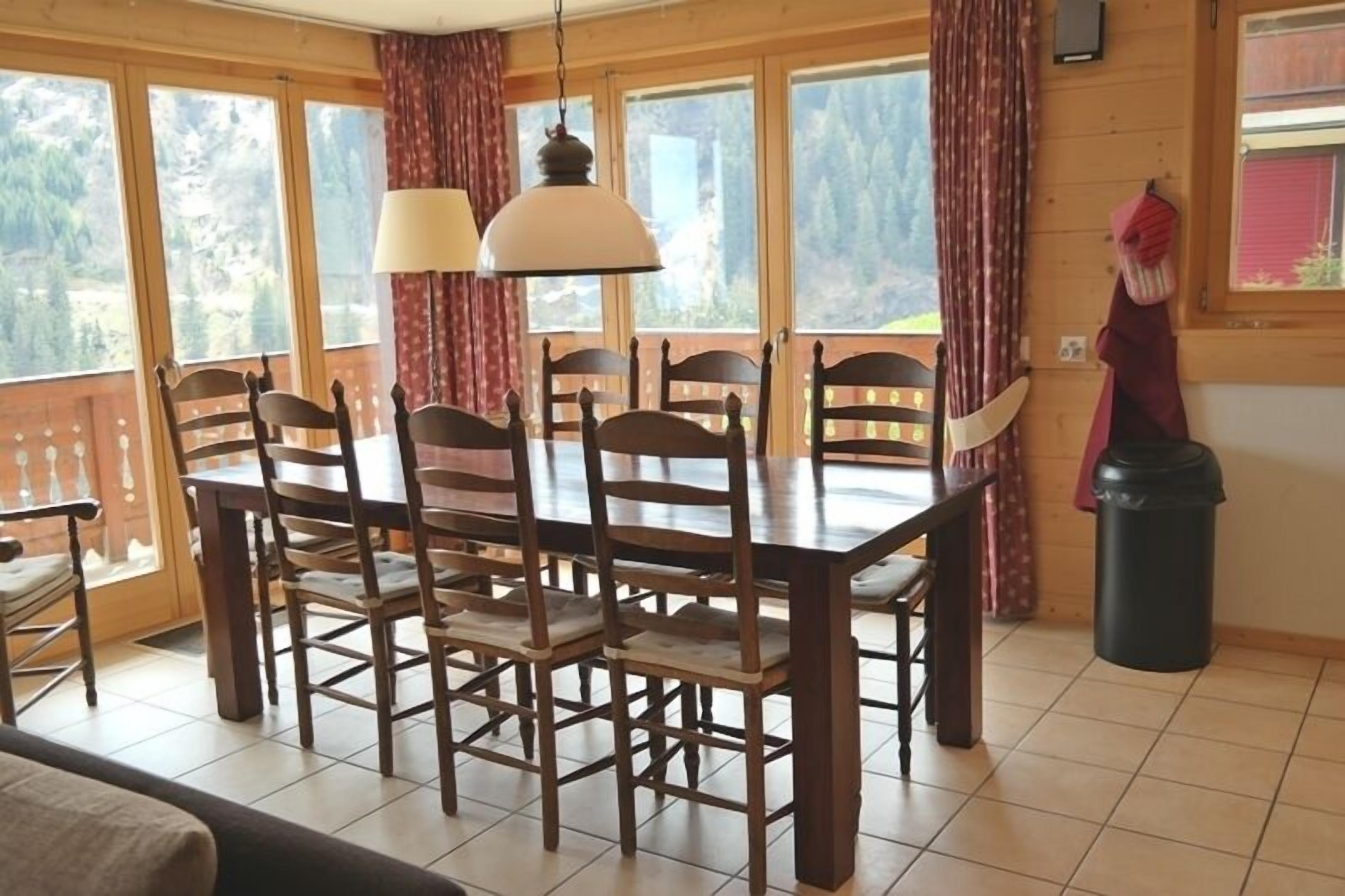 Une table à manger en bois, avec six chaises, de grandes fenêtres ornées de rideaux et une vue sur un paysage montagneux.