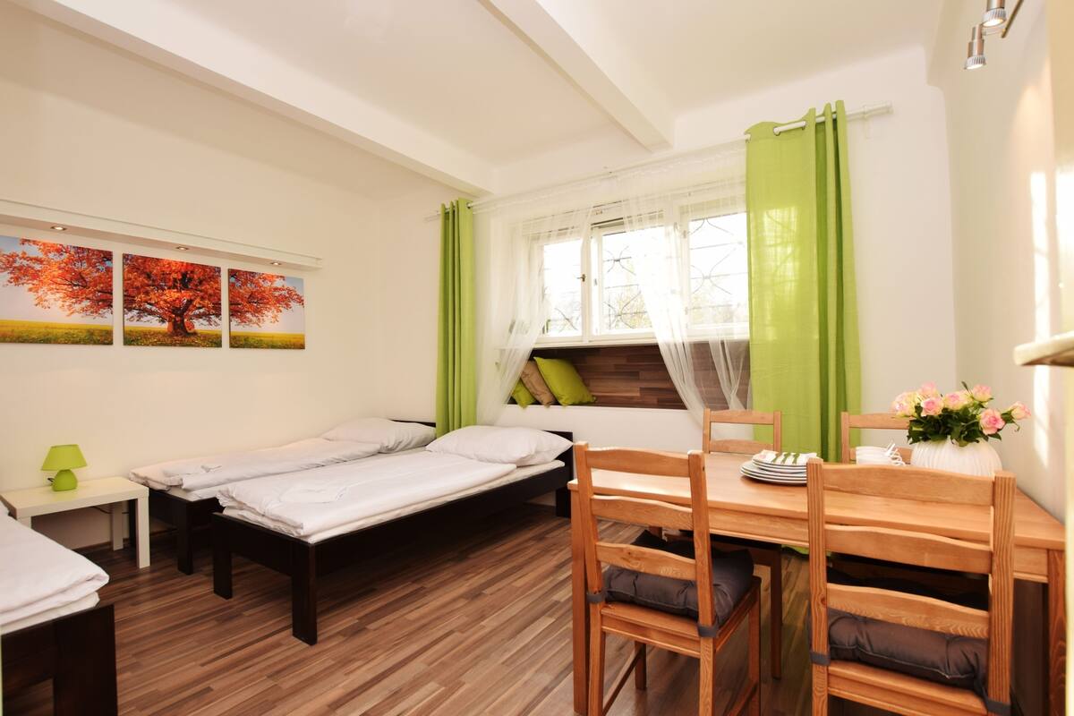 A bedroom with two single beds, a wooden dining table with chairs, a small bedside table with a lamp, and a window with green curtains.