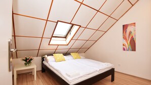 A bedroom with a sloped ceiling, a skylight, a bed with white linens, two yellow pillows, a bedside table with a vase of flowers, and a colorful abstract painting on the wall. - Císařka apartment (Prague)