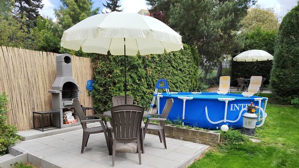 A patio with a table and chairs, a barbecue grill, a swimming pool, and a large umbrella. - Císařka apartment (Prague)