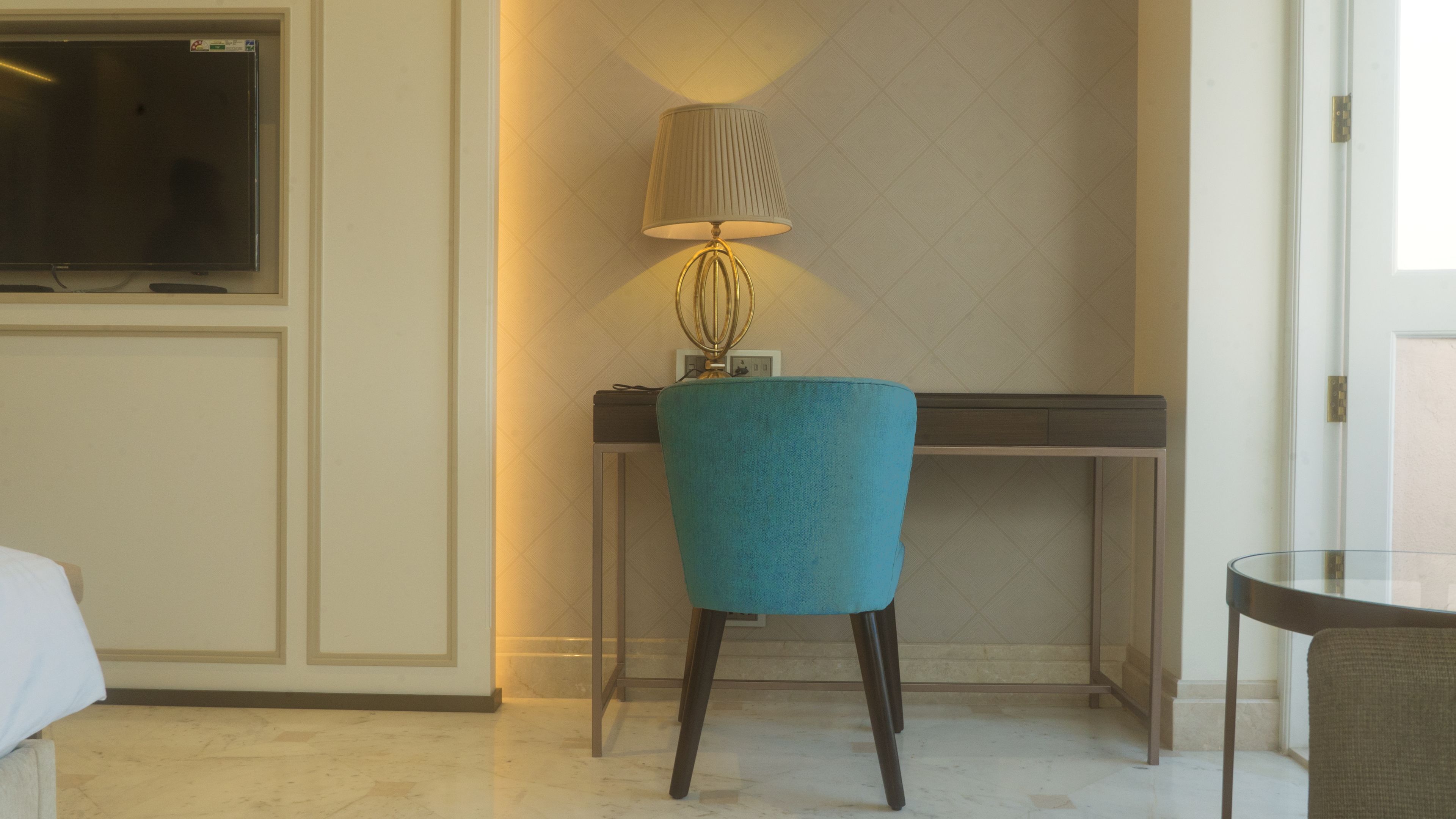 A hotel room with a teal chair, a desk, a lamp, and a TV.