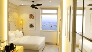 Une chambre d’hôtel avec un lit, un ventilateur de plafond, une fenêtre donnant sur le port et des œuvres d’art encadrées accrochées au mur.