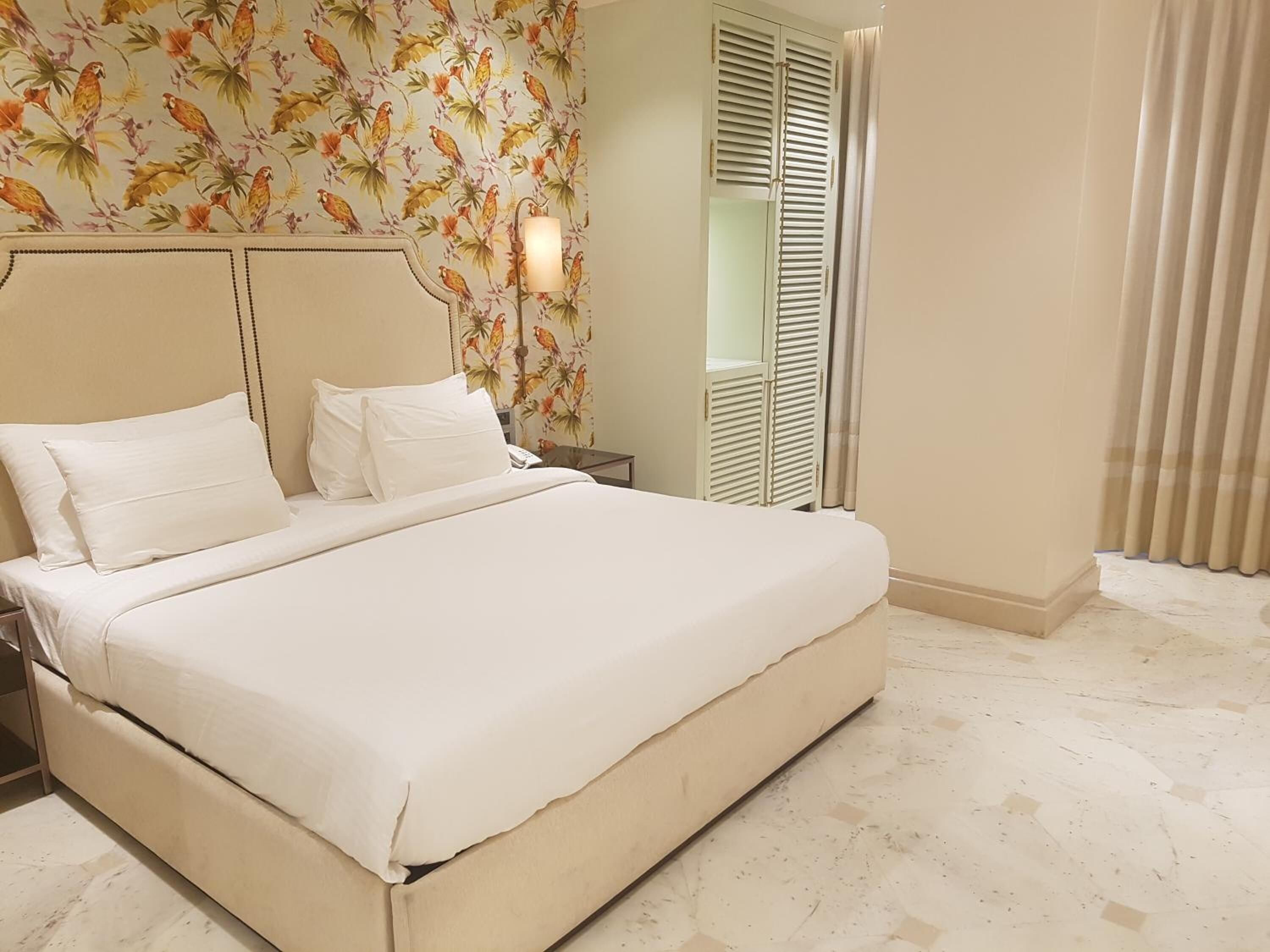 A hotel room with a large bed, a nightstand, a lamp, a wardrobe with louvered doors, and a floral wallpaper.