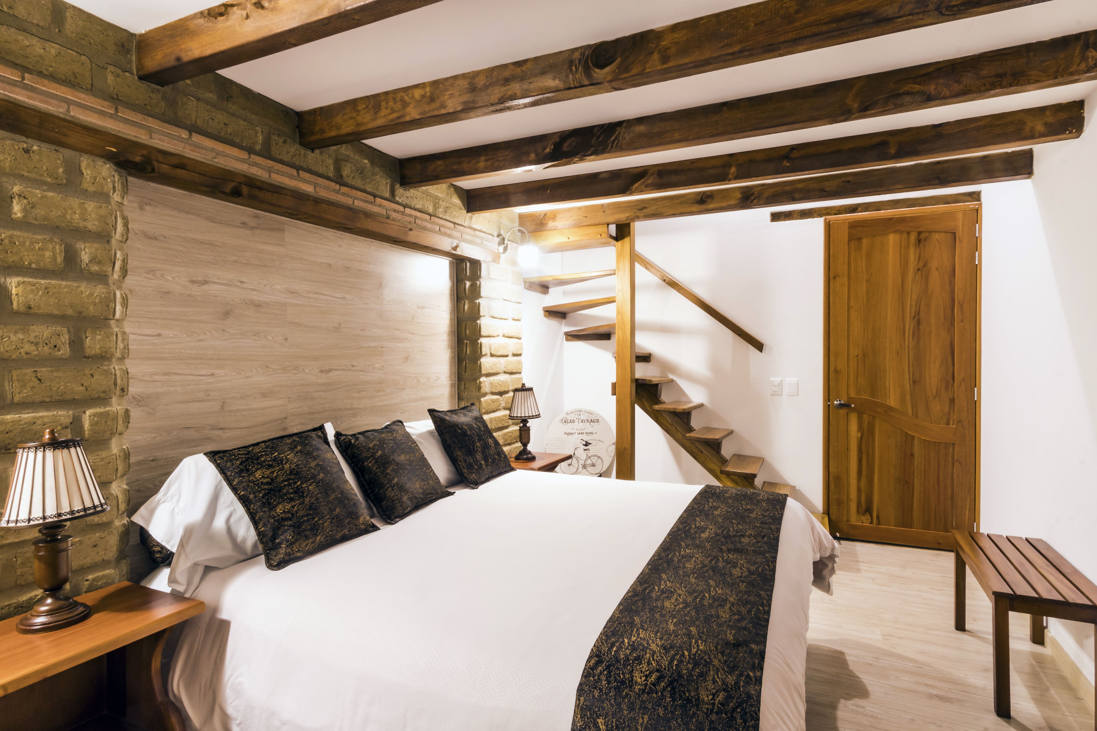 A bedroom with a large bed, wooden beams, a brick wall, a wooden door, and a staircase.