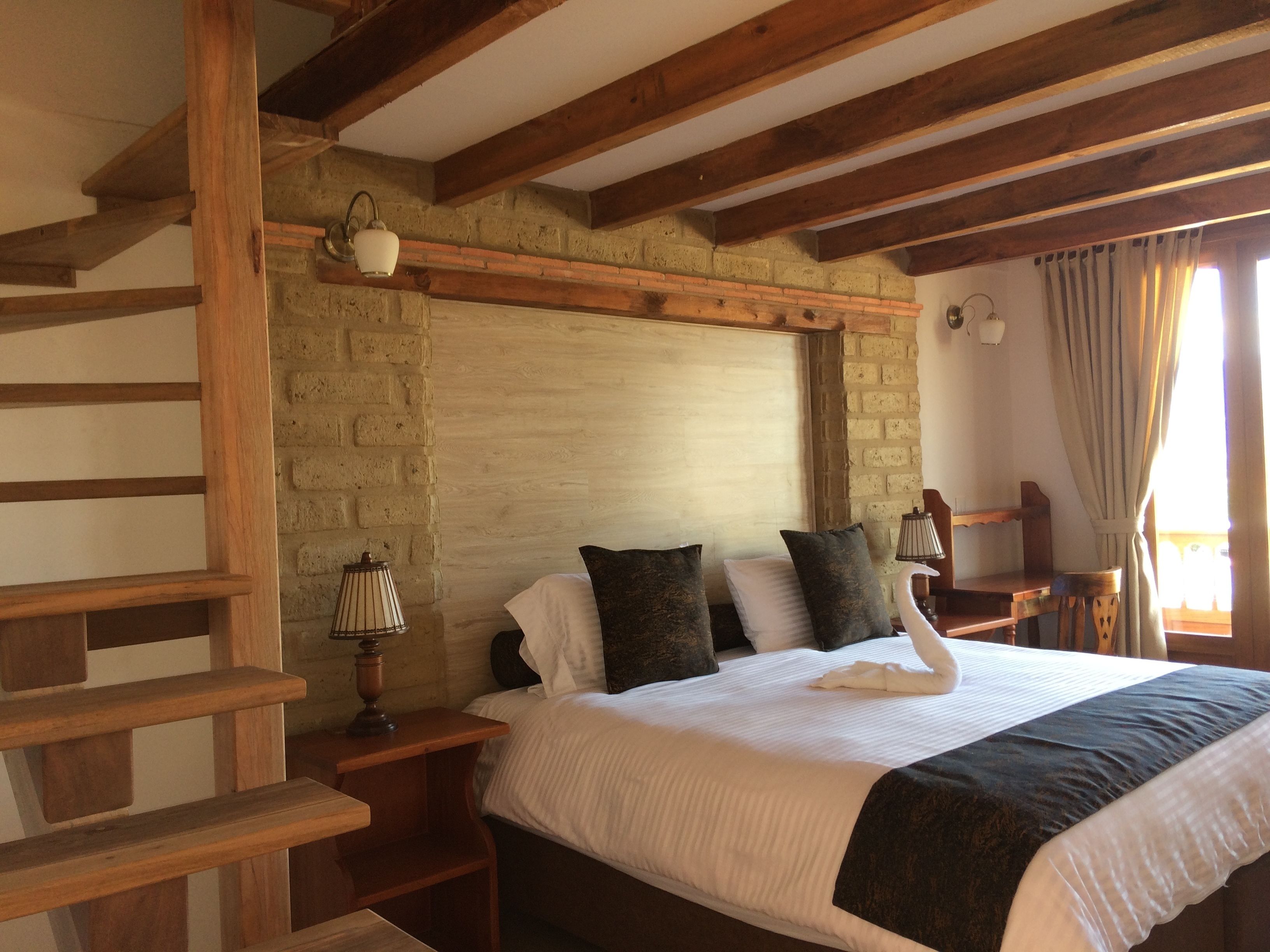 A bedroom with a wooden headboard, a brick wall, a bed with white and dark pillows, a nightstand with a lamp, and a wooden chair.
