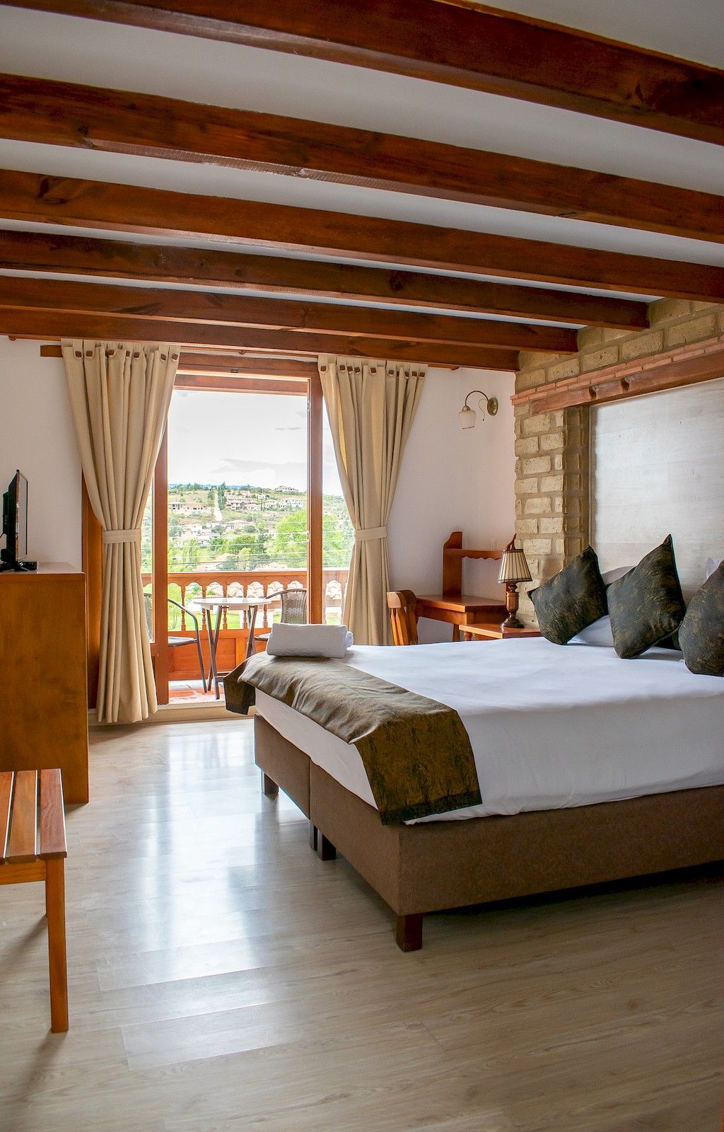 A bedroom with a large bed, wooden beams, a desk, a chair, and a view of greenery outside.