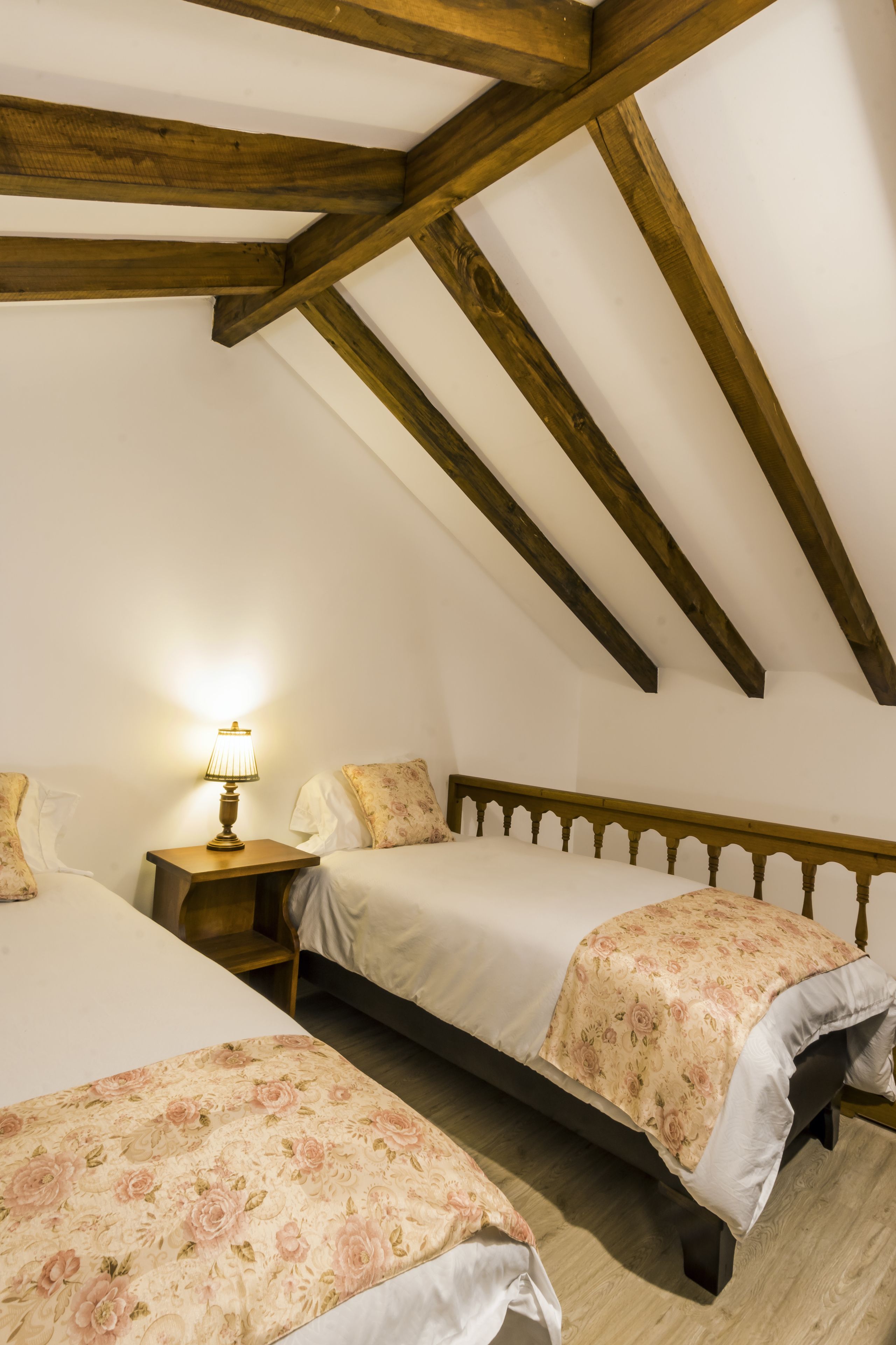 A bedroom with two beds, wooden beams on the ceiling, and a bedside table with a lamp.