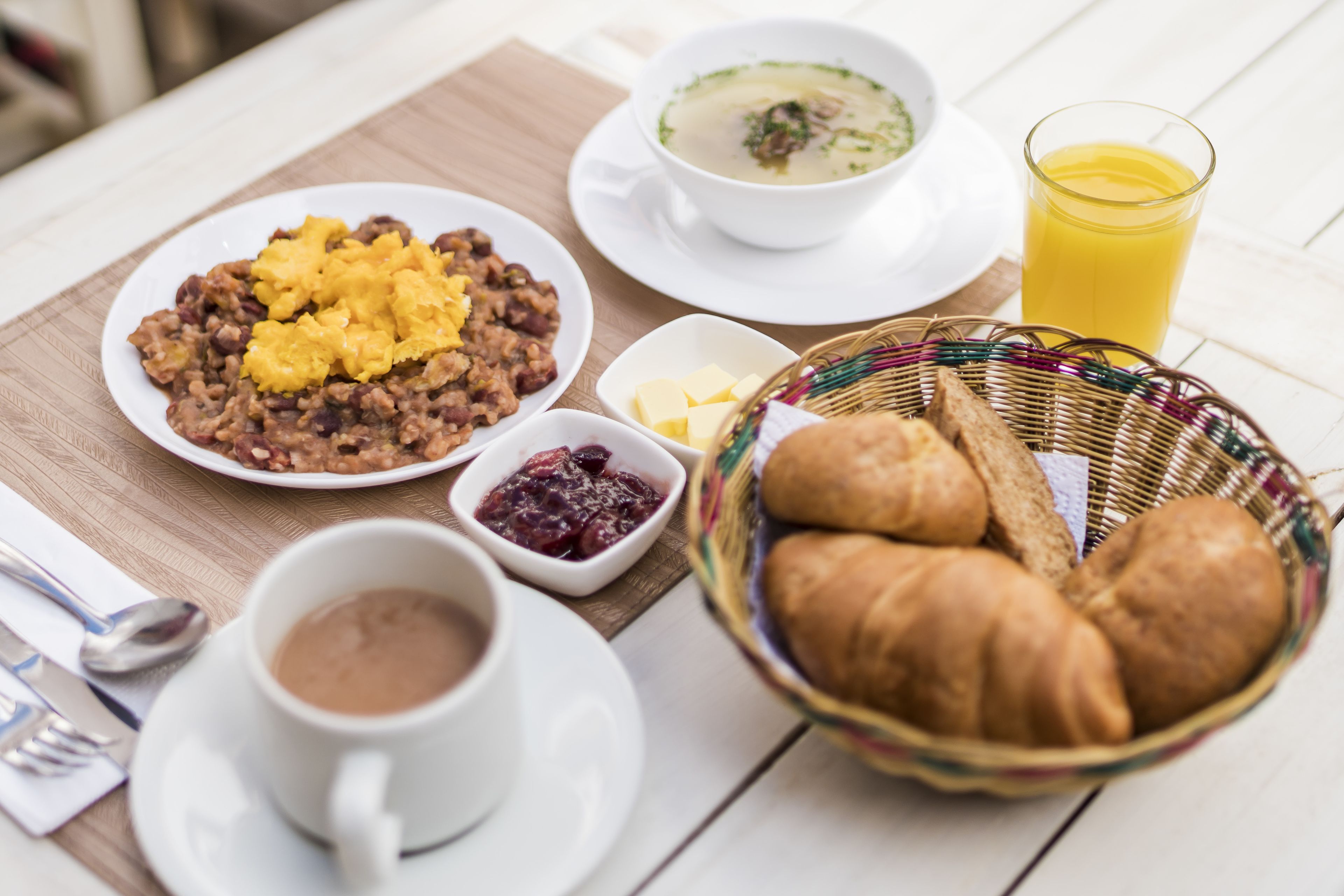 Un desayuno con un plato de frijoles revueltos y huevos, un tazón de sopa, un vaso de jugo de naranja, una cesta de cruasanes y una taza de café.