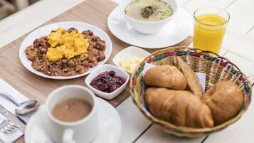 Un desayuno con un plato de frijoles revueltos y huevos, un tazón de sopa, un vaso de jugo de naranja, una cesta de cruasanes y una taza de café.