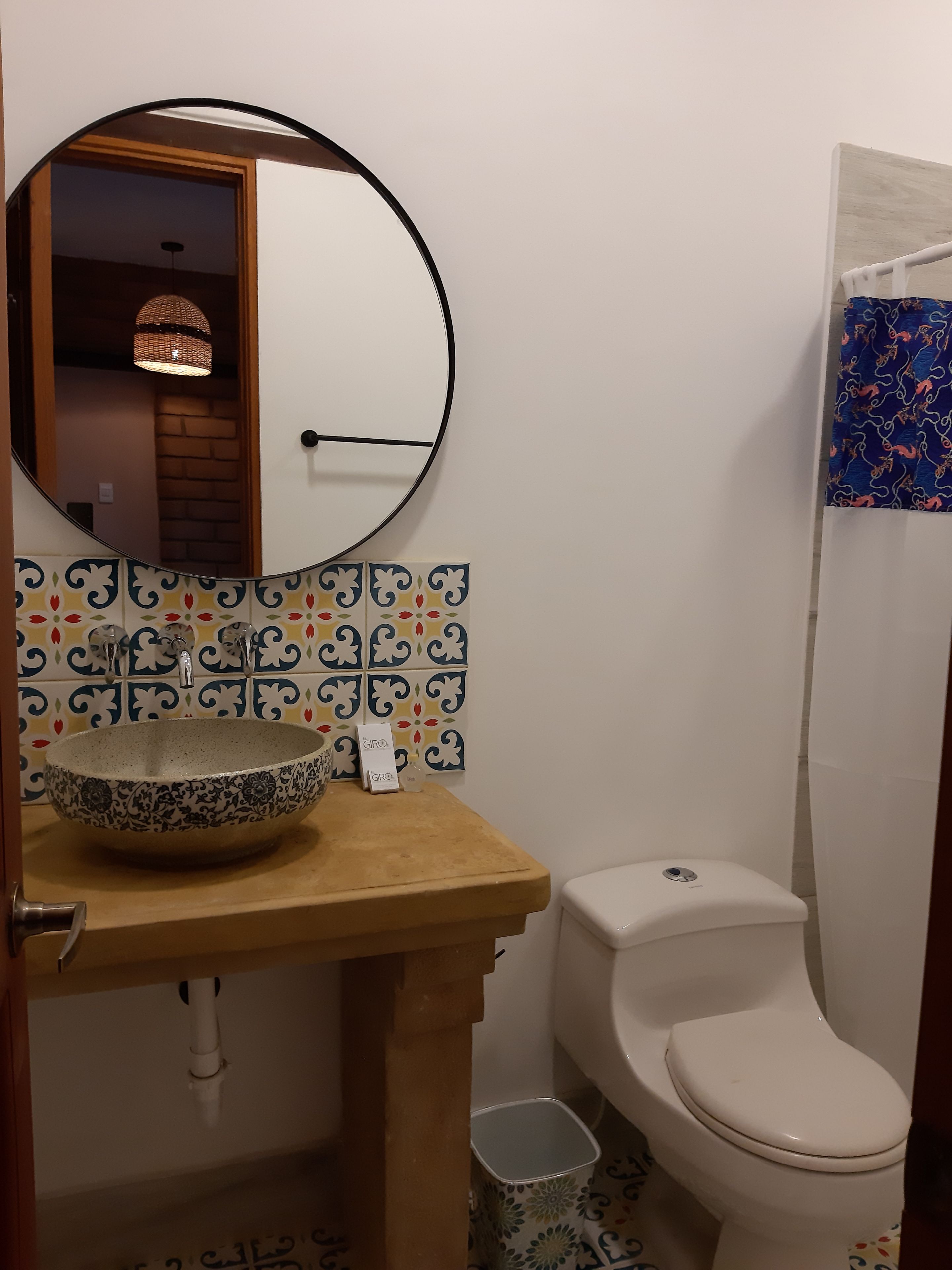 Une salle de bain avec un miroir rond, un lavabo agrémenté d’un récipient décoratif, des toilettes et un rideau de douche à motifs.