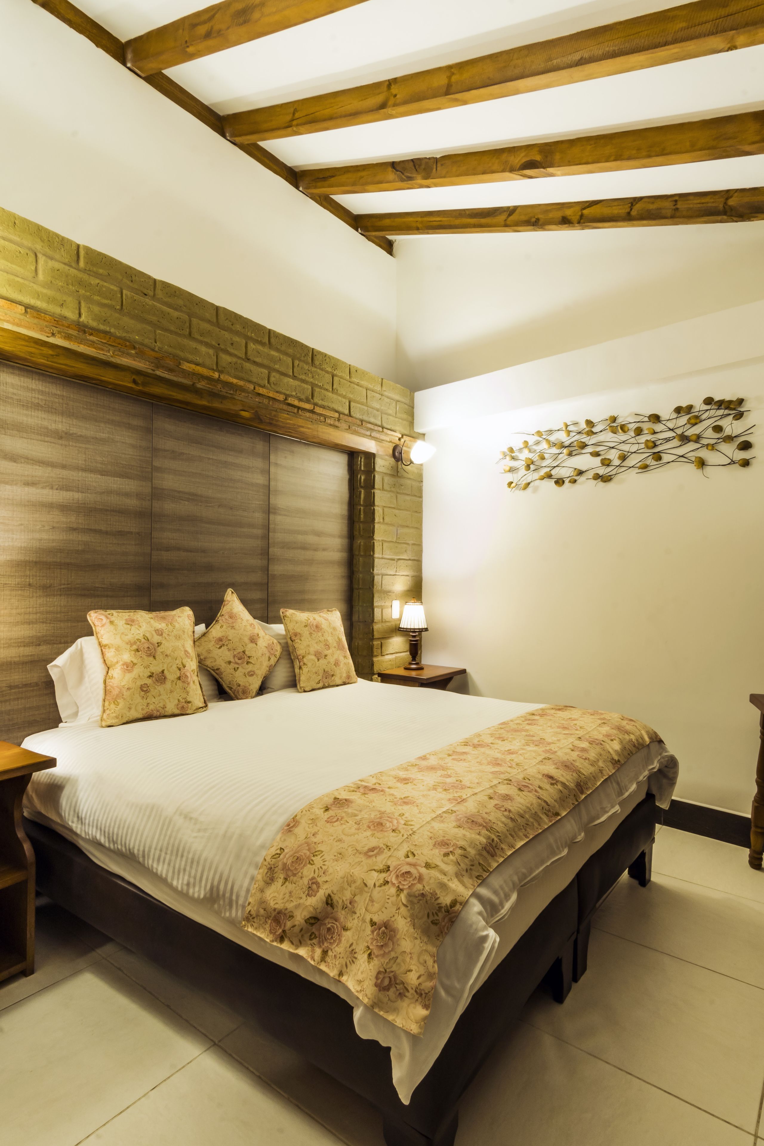 A bedroom with a wooden headboard, a large bed with floral bedding, a nightstand with a lamp, and a wall-mounted light fixture.