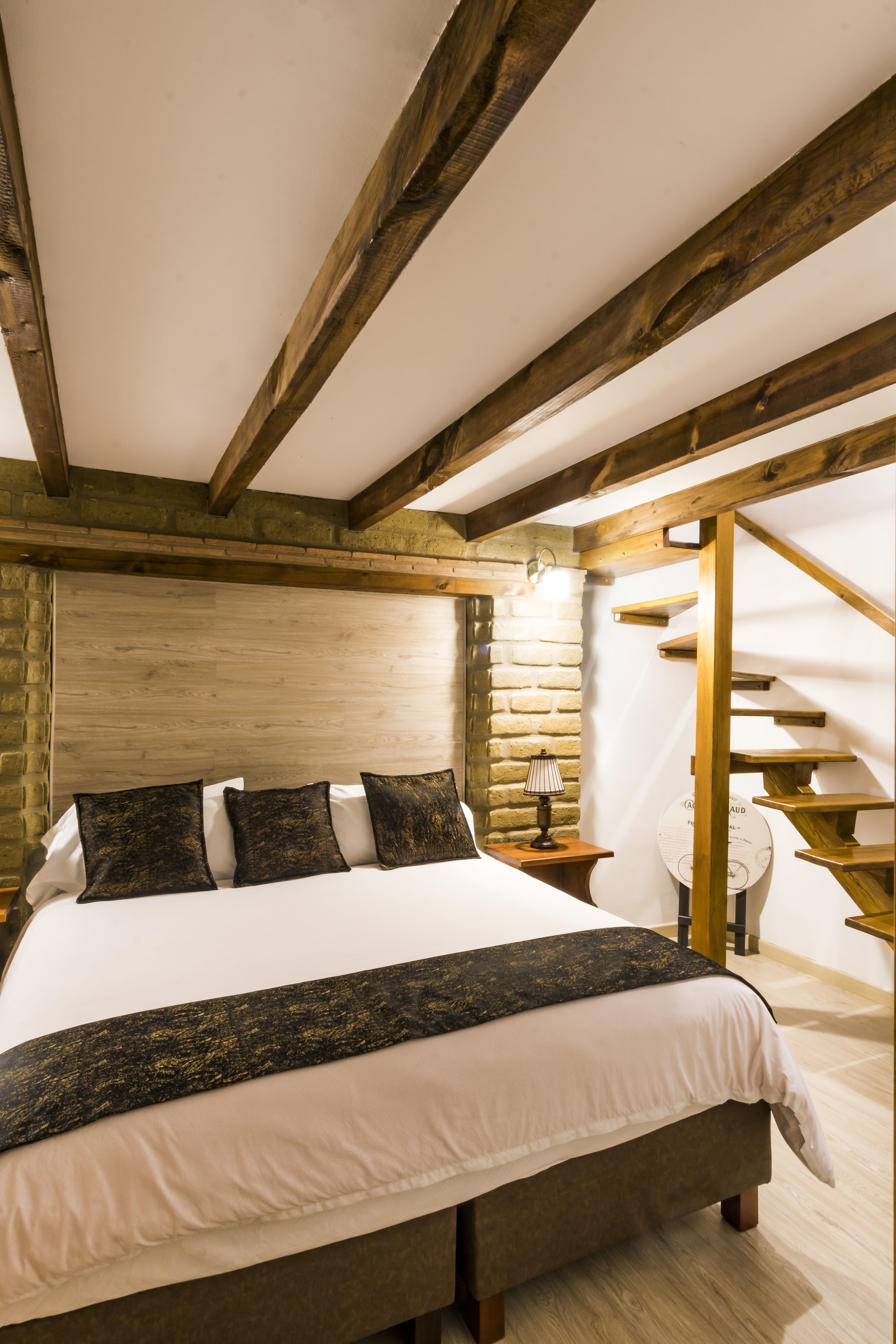 Un dormitorio con una cama grande, vigas de madera, una pared de piedra y una escalera.