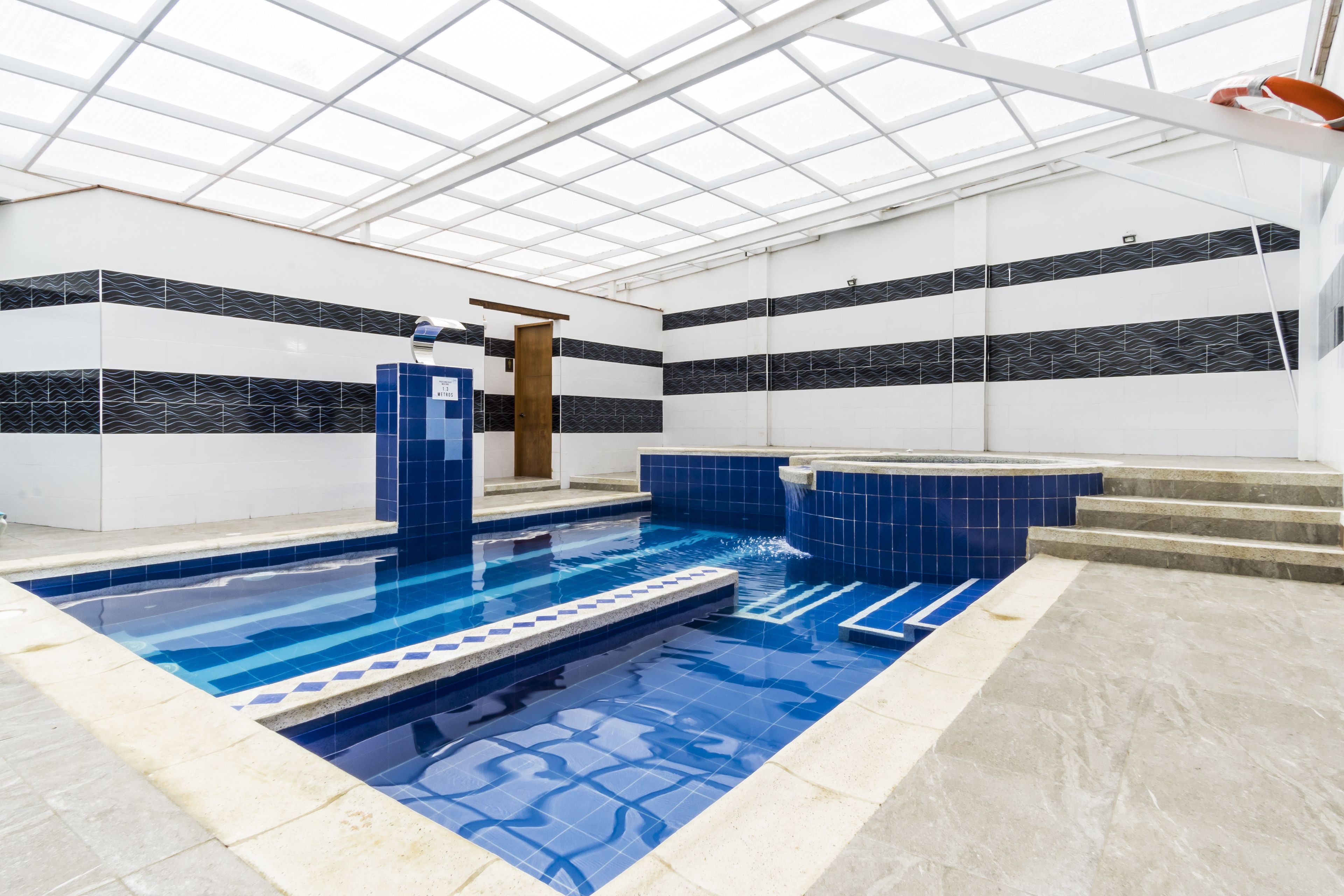 Una piscina interior con agua azul cristalina, paredes con azulejos y techo con tragaluces.