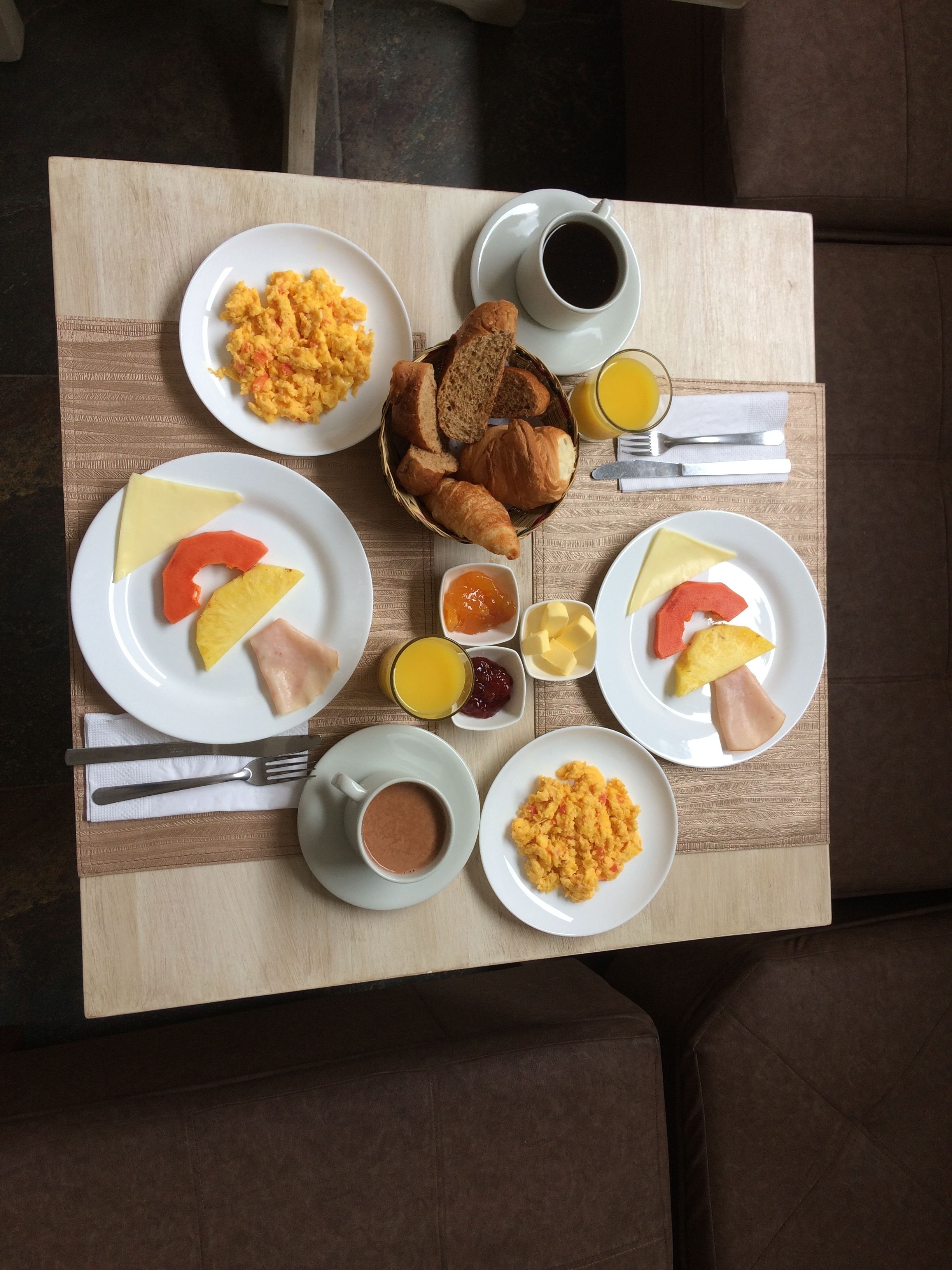 Una mesa de desayuno con huevos revueltos, tostadas y fruta, además de tazas de café y jugo de naranja.