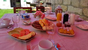 Un área de comedor al aire libre con un muro de piedra de fondo, mostrando una mesa dispuesta con alimentos para el desayuno, incluyendo pasteles, fruta y bebidas.