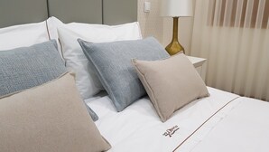 Una cama bien hecha con múltiples almohadas en tonos azules y beige, una lámpara de noche y un interruptor de luz en la pared.