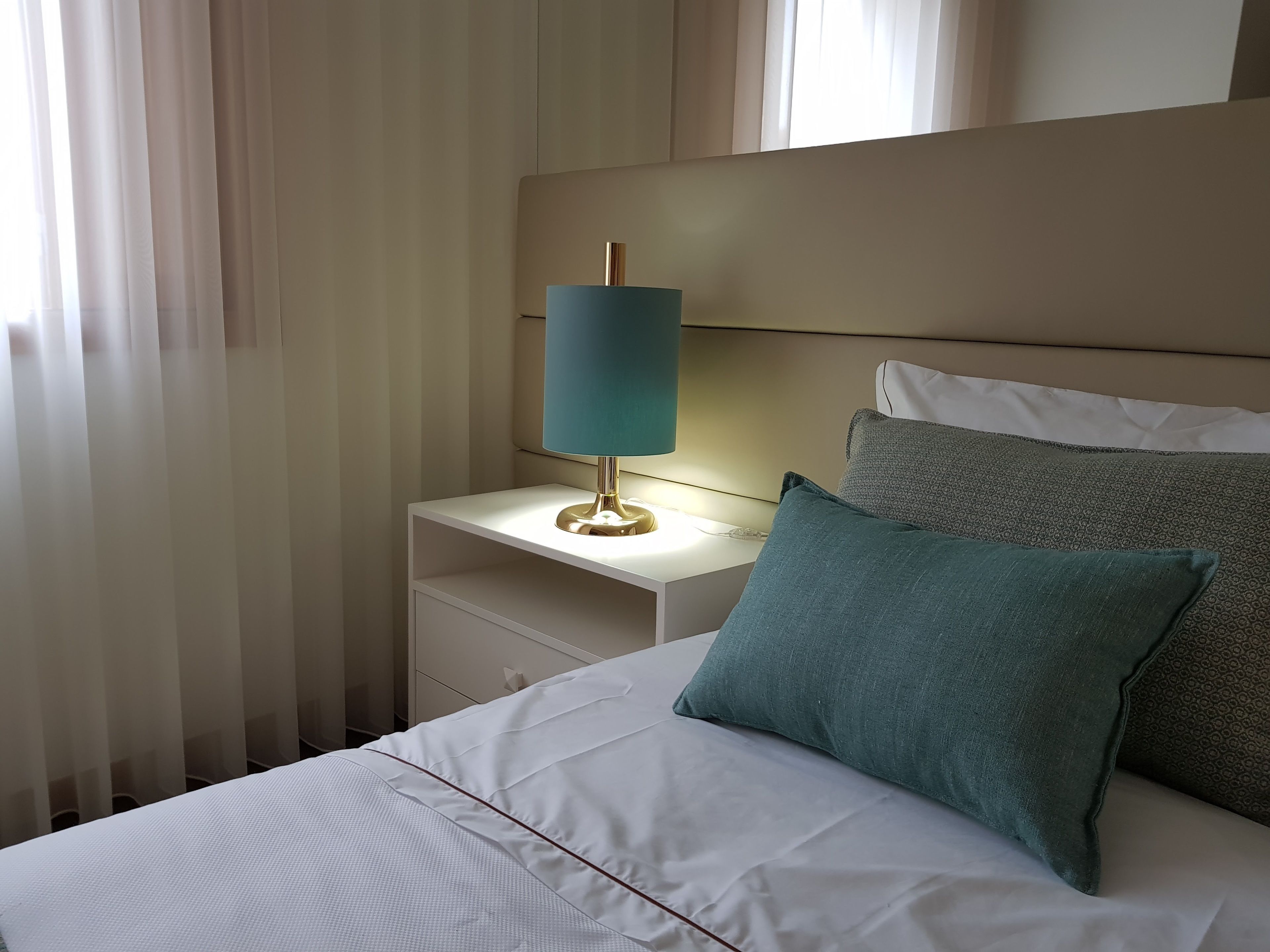 A neatly made bed with white bedding and teal pillows, a bedside table with a blue lamp, and sheer curtains.