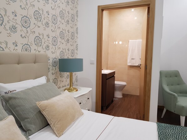 Un dormitorio ordenado con cama, mesita de noche, lámpara y un sillón verde.