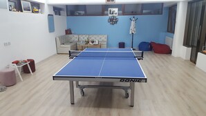 A room with a ping pong table, a sofa, a chair, and a small table with drinks.
