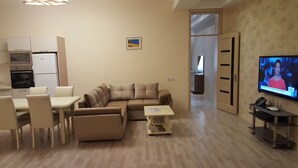 A modern living room with a beige sofa, a dining table with chairs, a TV, and a kitchen area with appliances.