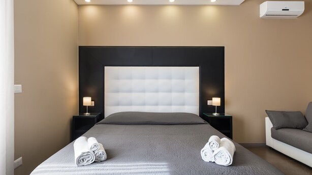 A modern hotel room with a large bed, two bedside tables with lamps, a grey sofa, and an air conditioning unit on the wall. - BH Napoli Railway (Naples)