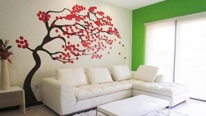 A modern living room with a white sectional sofa, a white coffee table, and a large wall mural of a tree with red flowers. - Heliko 102 by CocoBR (Playa del Carmen)