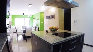 A modern kitchen with a granite countertop, a central island with a fruit bowl, and a dining area with chairs and a table. - Heliko 102 by CocoBR (Playa del Carmen)